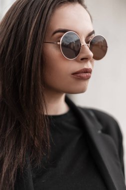 Şık bir portre. Kahverengi saçlı, seksi dudaklı, güzel, temiz tenli, şık güneş gözlüklü, modaya uygun, modaya uygun siyah ceketli, kentin sokaklarında beyaz duvarın yanında duran hoş bir kadın..