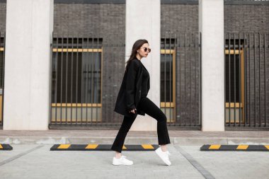 Moda güneş gözlüklü, modaya uygun siyah günlük kıyafetler içinde, beyaz deri ayakkabılı, havalı genç bir kadın modern binanın yakınında yürüyor. Gelişigüzel gençlik kıyafeti giyen çekici Avrupalı kız şehirde seyahat ediyor..