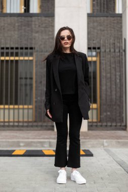 Modaya uygun, uzun ceketli, modaya uygun kot pantolonlu, şık tişörtlü ve modern binanın yakınında beyaz ayakkabılı güzel bir kadın. Moda bir bahar kıyafeti. Sıradan moda. Siyah biçim.