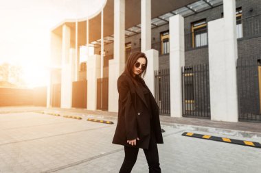 Moda mankeni, şık siyah elbiseli şık güneş gözlüklü genç bir kadın gün batımında caddeyi gezer. Dışarıda turuncu güneş ışığının arka planında güzel, modaya uygun bir kız. Bahar görünümü.