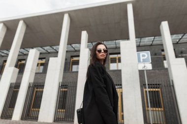 Modayı takip eden güneş gözlüklü, şık çantalı güzel bir genç bayan şehrin modern binasının yakınında yürüyor. Moda dünyasının güzel şehirli kızı sokakta günlük kıyafet giyiyor..