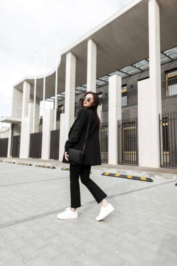 Moda desenli, deri çantalı, şık güneş gözlüklü, modaya uygun siyah elbiseli, modaya uygun genç koleksiyonu olan genç bir kadın şehrin modern binasının yanında yürüyor. Şehirli havalı kız sokakta poz verir.