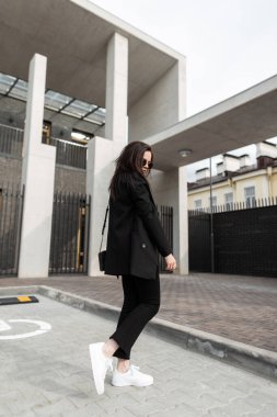 Eski moda güneş gözlüklü, modern binanın yakınında duran deri çantalı, şık, şık, genç bir bayan. Havalı, modaya uygun, siyah ceketli hippi kız.