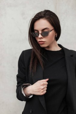 Şık bir portre, genç moda güneş gözlüklü, şık bir genç bayan, şehrin modern klasik duvarının yanında siyah, eski moda bir ceket giyiyor. Sokakta çekici Amerikalı manken kız.