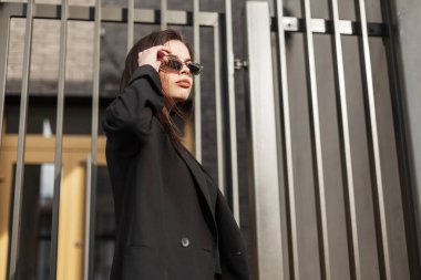 Eski moda güneş gözlüklü, siyah ceketli, seksi dudaklı modern genç kadın şehrin klasik duvarının yanında duruyor. Siyahlar içinde güzel havalı bir kız sokakta bardakları düzeltiyor..