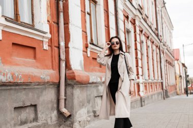 Moda güneş gözlüklü, şık bej renkli trençkotlu güzel bir kadın model şehrin klasik binasının yanında yürüyor. Güzel kız sokakta yürümekten hoşlanıyor. Kadınlar için bahar giysisi.