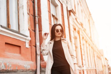 Şık güneş gözlüklü, modaya uygun siyah elbiseli bej rengi trençkotlu şık bir genç kadın gün batımında sokakta yürüyor. Seksi manken şehirde hafta sonu ve güneş ışığının tadını çıkarıyor..