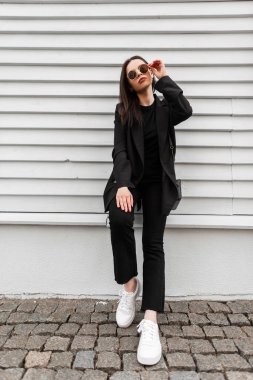 Beyaz, eski moda ayakkabılar içinde, el çantalı, şık siyah elbiseli güzel bir manken şehrin ahşap binasının yanında güneş gözlüğü takıyor. Sokakta poz veren sıradan elbiseli hoş bir kız..
