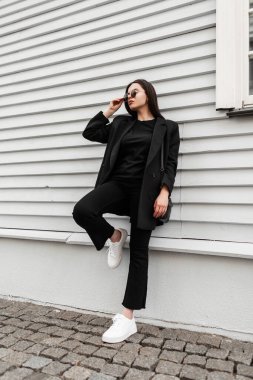 Spor ayakkabılı, spor ayakkabılı, güneş gözlüklü, spor ayakkabılı, spor ceketli muhteşem genç bayan caddedeki klasik ahşap binanın yanında dinleniyor. Güzel bir kız, açık havada şık bir gençlik kıyafeti giyiyor.