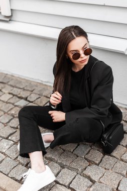 Yeni moda moda koleksiyonundan, şık güneş gözlüklü, şık siyah elbiseli güzel bir kadın kentteki eski ahşap binanın yanındaki taş fayansta oturuyor. Şehir kızı sokakta dinleniyor.