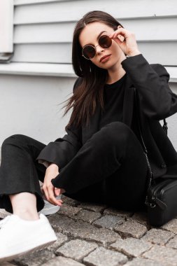Moda mankeni, şık güneş gözlüklü, modaya uygun siyah elbiseli, modaya uygun moda koleksiyonu olan genç bir kadın kentteki klasik ahşap binanın yanında dinleniyor. Güzel bir kız taş fayansların üzerinde oturuyor.