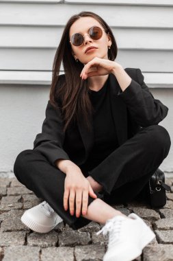 Şık, genç, seksi dudaklı, şık genç bayan hippi güneş gözlüklü, siyah elbiseli, beyaz renkli spor ayakkabılı, kentteki ahşap binanın yanında poz veriyor. Amerikalı manken kız sokaklarda dinleniyor..