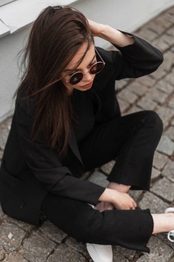 Havalı, modern, güneş gözlüklü, modaya uygun siyah giysili genç bir kadın şehirdeki klasik ahşap binanın yanında saç düzleştiriyor. Amerikalı modacı kız model taş fayansların üzerinde dinleniyor..