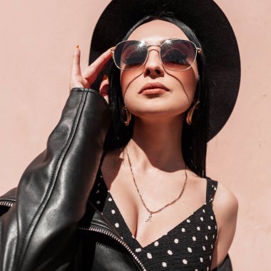 Güneşli bir portre, zarif bir şapkayla, şık bir elbiseyle, siyah deri ceketli, kumsalda pembe duvarın yanında elbisesi olan güzel esmer bir kadın. Moda kıyafetli seksi kız..