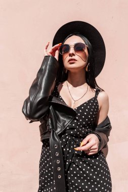 Moda çantalı, şık güneş gözlüklü, deri ceketli, güzel elbiseli çekici genç bir kadın güneşli bir günde şapkasını düzeltir. Zarif siyah yaz kıyafetleri içinde güzel moda bir kız açık havada güneş ışığının tadını çıkarıyor.