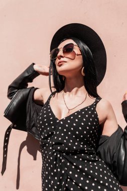 Moda gözlüklü, şapkalı, şık, siyah yazlık elbiseli seksi genç kadın pembe vintage duvarın yanındaki sokakta güneş ışığının tadını çıkarıyor. Güzel elbiseli çekici moda kız modeli güneşte duruyor..