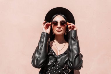 Avrupalı moda kadın güneşli bir günde pembe duvarın yanında güneş gözlüğü takıyor. Şık siyah elbiseli, şık şapkalı güzel bir kız binanın yanında poz veriyor. Güneş ışınları ve güzel bayan..