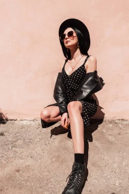 Moda elbisesi giyen, siyah şık şapkalı, klasik güneş gözlüklü, deri ceketli, şık genç bir kadın, sokakta pembe duvarın yanında oturuyor. Moda kıyafetleri içinde güzel bir manken yaz güneşli günlerinde rahatlıyor.