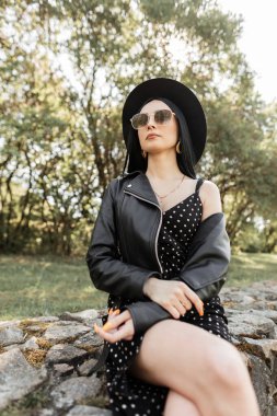 Güneşli bir günde moda güneş gözlüklü, zarif şapkalı, modaya uygun siyah yaz kıyafetleriyle güzel bir kadın portresi. Moda kıyafeti giymiş şık bir kız parkta taşların üzerinde oturuyor. Kadınlar için bahar kıyafeti