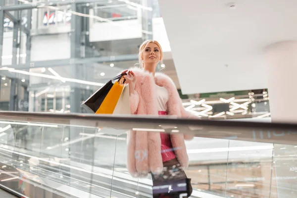 Modern mağazadaki yürüyen merdivende deri etekli, pembe kürklü, neşeli, genç bir manken kadın. Alışveriş torbalı mutlu kız alışveriş merkezinde seyahat ediyor. Bayan alışverişkolik..