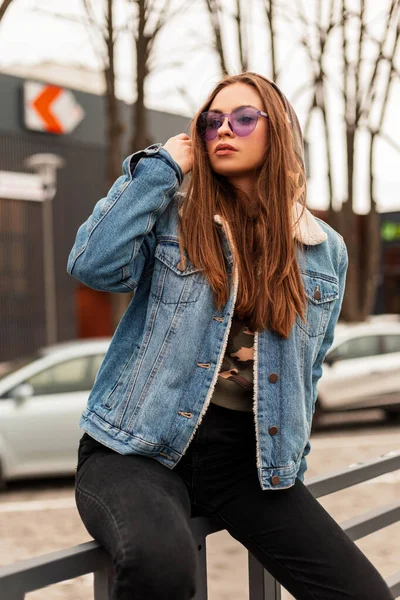 ファッショナブルな特大ブルーデニムジャケットで流行の紫色のメガネでスタイリッシュなモデルの若いヒップスター女性は 通りの駐車場の近くに長い髪をまっすぐにします 現代の女の子ヒップスターでヴィンテージ服で街 — ストック写真