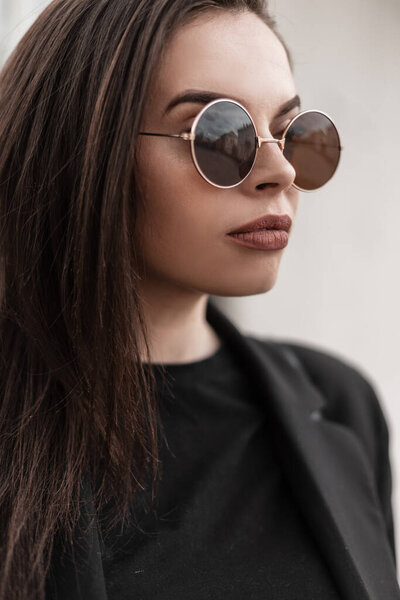 Stylish portrait pretty young lovely woman with brown hair with sexy lips with beautiful clean skin in fashionable cool sunglasses in black trendy blazer near vintage white wall on street in city.