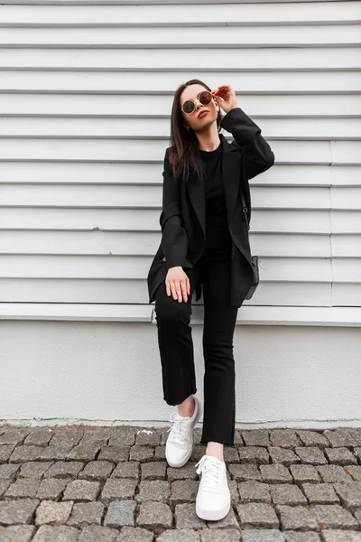 ハンドバッグとスタイリッシュな黒の服の白いヴィンテージの靴のかなり若い女性のファッションモデルは 市内の木造建築物の近くのファッションサングラスをまっすぐにします ファイン女の子でカジュアルドレスで通りにポーズ — ストック写真