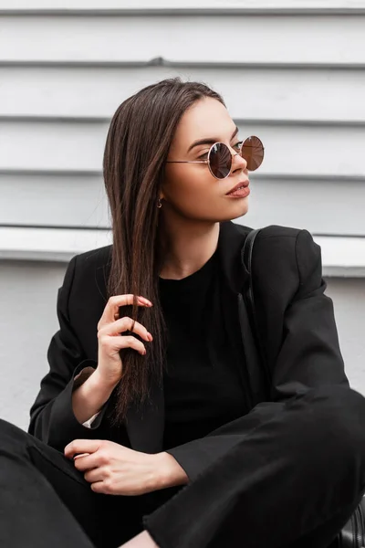 流行の若い黒の服でスタイリッシュなサングラスで流行の若い女性のヒップスターは 屋外で木製のグレーのヴィンテージの壁の近くに座っている 魅力的なかわいいです豪華な女の子ファッションモデルリラックスオンストリート — ストック写真