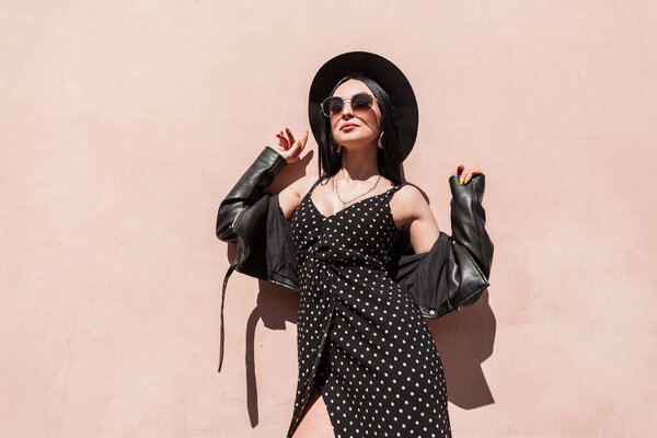 Stylish young woman model in hat in jacket in dress in sunglasses enjoys sun in city. Fashion portrait vogue girl in black elegant summer beautiful clothes in bright sunlight near vintage pink wall.