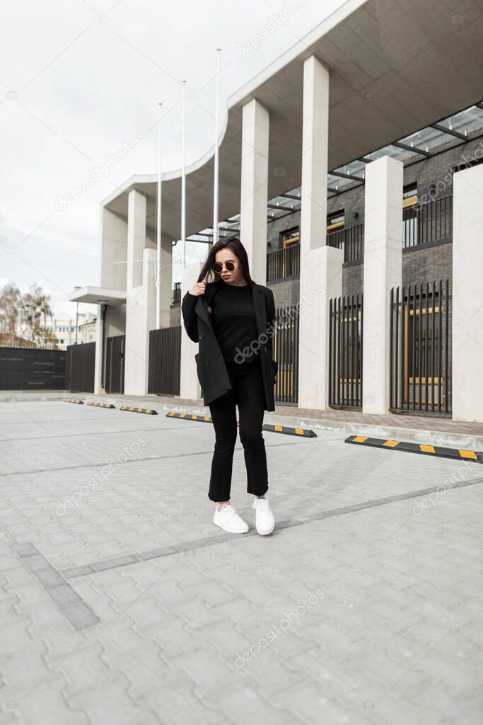 Fashion model attractive young woman in stylish sunglasses in fashionable black clothes from new summer trendy collection is walks near modern building in city. Fine girl in vintage jacket outdoors.