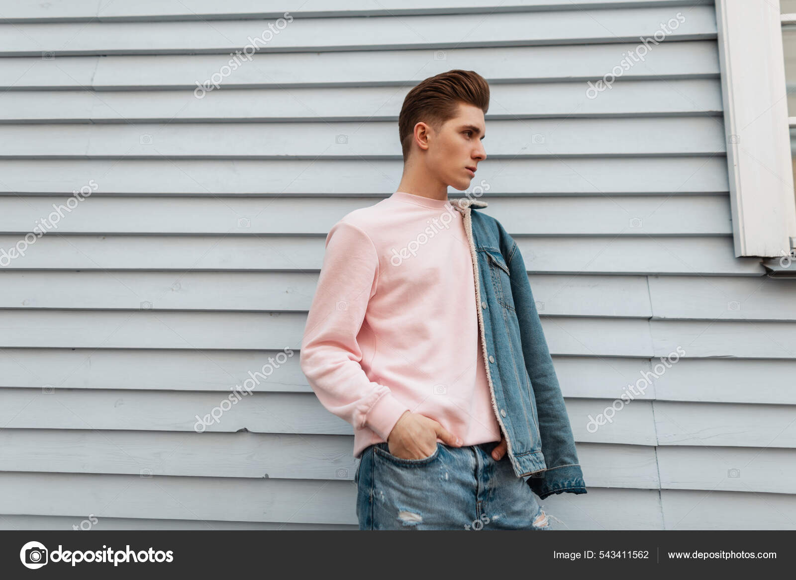 Hombre joven con estilo en sudadera rosa de moda en jeans de moda azul en  zapatillas blancas se sienta en baldosas de piedra cerca de edificio  vintage en la calle en la