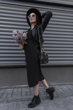 Şık siyah elbiseli, modaya uygun şapkalı, şık güneş gözlüklü, içinde buket desenli leylak çiçekleri olan şık bir kadın sokakta gri metal duvarın yanında duruyor. Bahar siyahı kıyafetli güzel kız..