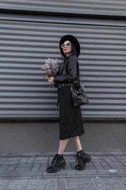 Şık şapkalı, güneş gözlüklü, siyah elbiseli, açık havada metal duvarın yanında duran leylak çiçekli şık kadın esmer. Tarz sahibi kız yürümekten ve taze güzel çiçeklerden hoşlanır..