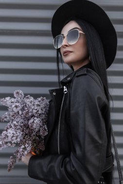Moda şapkalı, güneş gözlüklü, şık deri ceketli, kentin klasik metal duvarının yanında menekşe çiçekli güzel bir kadın portresi. Leylak bitkileriyle genç ve güzel bir kız.