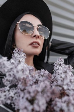 Taze portre çekici esmer kadın, lüks moda şapkalı, siyah ceketli, güneş gözlüklü, kentin metal duvarının yanında güzel leylak çiçekleri olan. Seksi kız modeli ve güzel bitkiler..