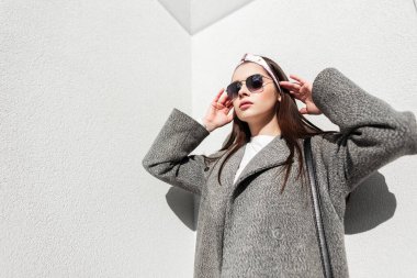 Şık bahar kıyafetleri içindeki çekici genç kadın güneşli bir günde duvarın yanındaki saçları düzleştiriyor. Parlak güneşli bir günde bandanalı güneş gözlüklü şık bir kız şehirde poz veriyor. Gençlik modası.