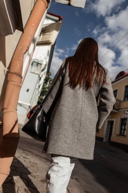 Şık giyinmiş, kot pantolonlu ve çantalı şık bir kadın güneşli bir günde sokakta yürüyor.