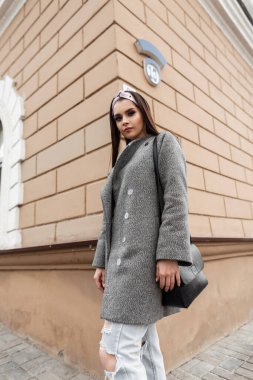 Şık giyinmiş, şık bir kadın mankeni, ceket, kot pantolon ve siyah bir çanta şehirde klasik bir binanın yanında yürüyor.