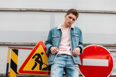Moda mankeni, şık mavi kot elbiseli, pembe tişörtlü yakışıklı genç adam şehirde metal yol işaretlerinin yanında eğleniyor. Günlük kot elbiseli modacı. Sokak gündelik bahar-yaz tarzı.