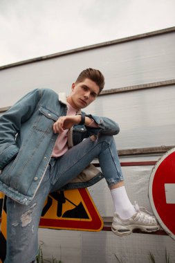 Mavi gençlik gündelik kotlu, pembe tişörtlü, ciddi bir genç adam kentteki caddede metal yol tabelasının yanında duruyor. Havalı kot giyimli, modaya uygun bir adam modeli. Erkek giyim