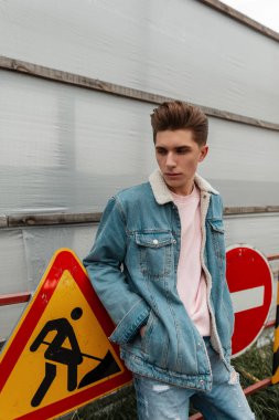 Pembe şık tişörtlü, şık mavi kot ceketli yakışıklı genç adam caddedeki metal yol işaretlerinin yanında duruyor. Açık havada poz veren modaya uygun genç kotları olan çekici bir adam. Sıradan erkek giyim tarzı.