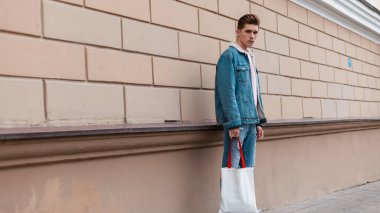 Şık gençlik kıyafetleri içinde şık bir genç adam. Göz kamaştırıcı pembe tişörtler içinde, kumaş çanta yanında duruyor. Moda kıyafetleri içinde, sokak kıyafetleri içinde şık bir adam modeli..