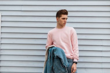 Pembe şık kazaklı, kotlu mavi ceketli yakışıklı genç adam caddedeki beyaz vintage binanın yanında duruyor. Yakışıklı, modacı, şehirde saç stili olan bir model.. 