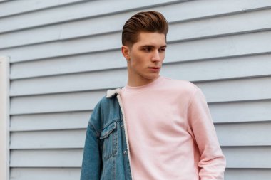 Moda kot pantolonlu, mavi ceketli, açık havada beyaz duvarın yanında poz veren yakışıklı genç adam modeli. Yeni koleksiyon pozlarından şık kotlar giyen şehirli adam sokakta poz veriyor..