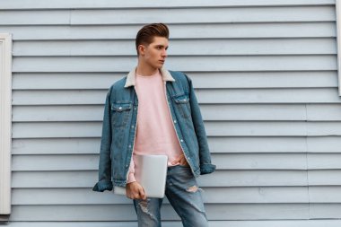 Şık saç stili, modaya uygun mavi kot pantolonları ve modern aletleri olan yakışıklı genç adam şehrin klasik binasının yanında duruyor. Moda mankeni, laptoplu çekici adam sokakta poz veriyor.