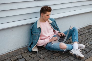 Başarılı, genç ve çekici adam dizüstü bilgisayarlı şık kot elbiseli serbest çalışan sokaktaki klasik binanın yanındaki taş fayansta oturuyor. Moda tasarımcısı yakışıklı adam açık havada çalışıyor..