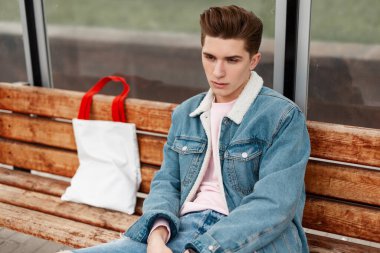 Şık kot ceketli, pembe tişörtlü, antika kotlu, ahşap bankta kumaş alışverişi yapan, şehirdeki kamu binasının önünde duran genç bir portre. Havalı elbiseli çekici adam dışarıda dinleniyor..