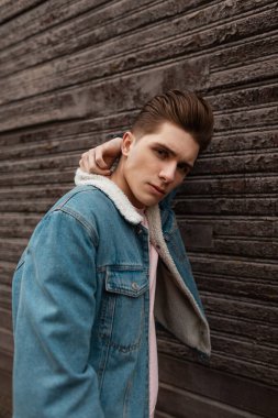 Modaya uygun kot ceketli, açık havada eski kahverengi ahşap duvarın yanında poz veren tatlı seksi genç adam mankeni. Yeni koleksiyondaki kotlu adam sokakta poz veriyor. Gençlik tarzı.