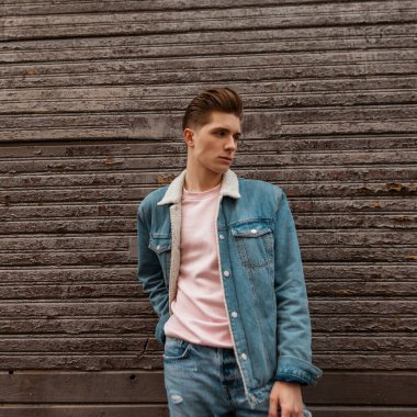 Ciddi yüzlü, modaya uygun saç stili, pembe tişörtlü, modaya uygun mavi kot pantolon giyen genç hoş bir adam sokaktaki eski ahşap duvarın yanında poz veriyor. Moda gençlik kotu giyen erkek manken dinlenmenin keyfini çıkarıyor..