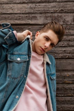 Pembe tişörtlü Avrupalı genç adam sokaktaki klasik ahşap duvarın yanında şık kot ceket düzeltiyor. Sıradan moda kotları içinde yakışıklı bir adam. Moda portresi. Genç erkek giyim..